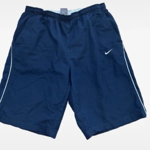 shorts från Nike - Snygga marinblå shorts från Nike med vit logga och vita detaljer längs sidorna. Shortsen har elastisk midja och fickor både fram och bak, perfekta för en sportig look.