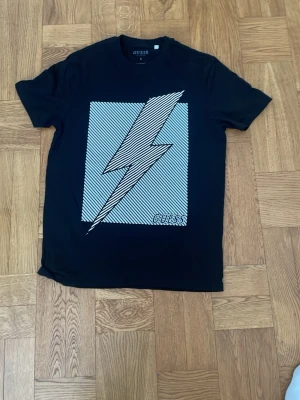 Svart t-shirt med blixt från Guess - Svart t-shirt från Guess med ett stort blixtmotiv i vitt och tunna ränder på bröstet. Klassisk rund hals och korta ärmar. 