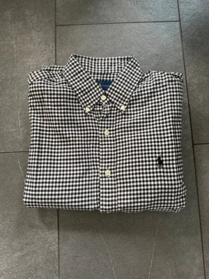 Rutig skjorta från Ralph Lauren - Klassisk svartvit rutig skjorta från Ralph Lauren med button-down krage och broderad logga på bröstet. Skjortan har långa ärmar, vita knappar och är tillverkad i mjuk bomull. Perfekt för en stilren och avslappnad look.