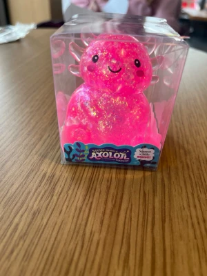 Glittrig rosa Axolotl Squeezy - En söt och glittrig rosa Axolotl Squeezy i mjukt material, perfekt som stressleksak eller dekoration. Kommer i en genomskinlig plastförpackning och har ett gulligt ansikte med svarta ögon och leende. Perfekt för dig som gillar roliga och färgglada prylar. Oöppnad köpt för 80kr 