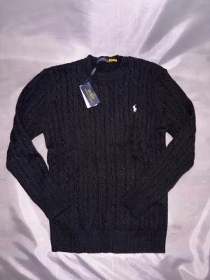Svart kabelstickad tröja Polo Ralph Lauren - Kabelstickad svart tröja från Polo Ralph Lauren med rund halsringning och den klassiska vita logobroderingen på bröstet. Tröjan har långa ärmar och ribbade muddar vid ärmslut och nederkant. Perfekt för en stilren och tidlös look.