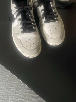 Air Jordan vita sneakers med svart detalj - Säljer ett par vita Air Jordan sneakers med svarta skosnören och svart detalj runt ankeln. Skorna har perforerad tå och röd Air Jordan-logga på hälen. Klassisk design i läder som passar dig som gillar streetstyle.