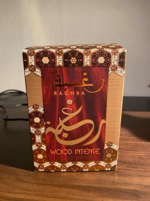 Raghba Wood Intense Eau de Parfum - Raghba Wood Intense Eau de Parfum med en lyxig, mörkbrun och guldig förpackning. Flaskan är genomskinlig med svart och gulddetaljer, och innehåller en bärnstensfärgad parfym. Doften har träiga och orientaliska inslag, perfekt för dig som gillar kraftfulla och unika parfymer.