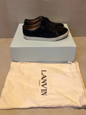 Mörkblå Lanvin sneakers med lackdetalj - Snygga svarta sneakers från Lanvin med ovandel i mocka och tåparti i blankt lack. Skorna har vita sulor och svarta skosnören. Insidan är fodrad i beige läder med Lanvin-logga. Perfekta för dig som gillar stilrena och exklusiva sneakers.