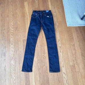 Mörkblå jeans från Nudie Jeans - Snygga mörkblå jeans från Nudie Jeans med klassisk femficksdesign och kontrastsömmar. Modellen har smal passform och ikoniska broderade bakfickor. Perfekta för dig som gillar stilrena och tidlösa jeans i 100% bomull. Modellen på jeansen är Thin finn. W28 L32