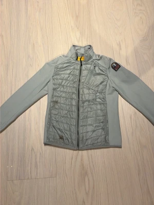 Ljusgrå pufferjacka från Parajumpers - Ljusgrå pufferjacka från Parajumpers med quiltad front, meshpaneler på sidorna och ärmarna, samt en gul detalj vid kragen. Jackan har en bröstficka med dragkedja och ett snyggt märke på ena ärmen. Perfekt för kalla vinterdagar.
