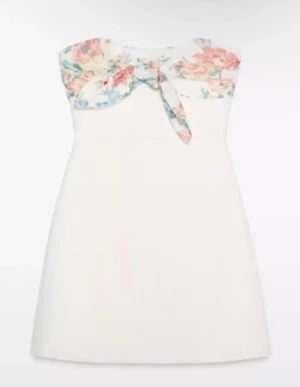 Vit kort klänning med blommig rosett - Supersöt vit kort klänning med blommigt mönster och stor rosett upptill. Axelbandslös design som ger en fräsch och somrig vibe. Perfekt för varma dagar och har en feminin passform. Materialet känns lätt och luftigt.