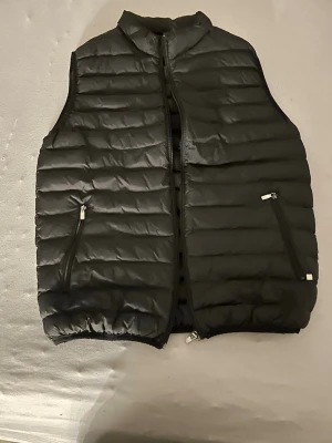Svart dunväst från hugo boss med dragkedjor - Stilren svart dunväst från hugo boss med horisontella sömmar och två praktiska fickor med dragkedja framtill. Västen har hög krage och stängs med dragkedja. Perfekt för lager-på-lager och enkel att matcha med olika outfits. Har sytt lite där bak 