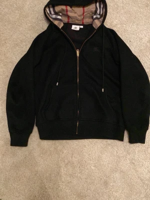 Svart hoodie från Burberry med dragkedja - Svart hoodie från Burberry med klassiskt rutigt foder i huvan och dragkedja framtill. Stora fickor på sidorna och justerbar huva med snören. Perfekt för en stilren och lyxig streetwear-look.