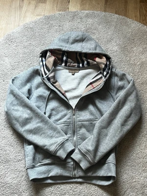 Grå zip-hoodie från Burberry S-M - Snygg grå hoodie med dragkedja från Burberry. Klassisk huva med det ikoniska rutmönstret på insidan, två fickor fram och ribbade muddar. Perfekt för en chill och stilren look. Mjuk bomullsblandning som känns skön mot huden.