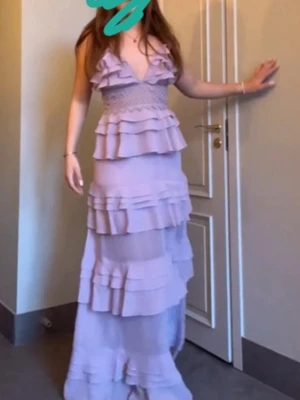 Ljuslila långklänning med volanger - Säljer en ljuslila långklänning med flera lager volanger och v-ringad front. Klänningen har smockad midja och är ärmlös, vilket ger en elegant och luftig känsla. Perfekt för dig som vill sticka ut med en romantisk look.