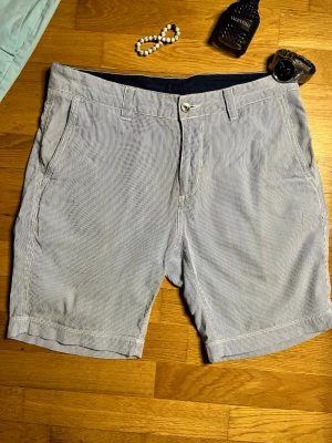 Randiga Bowman Chino shorts Sail Racing - Säljer ett par snygga randiga Bowman Chino shorts från Sail Racing. De är blå- och vitrandiga, har normal passform och är tillverkade i 100% bomull. Klassisk chino-modell med fickor fram och bak, perfekta för sommaren.