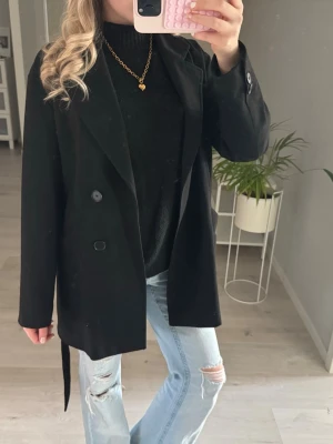Svart kappa  - Stilren svart kavaj med dubbelknäppning och tillhörande knytskärp i midjan. Klassisk krage och raka ärmar. Perfekt för en trendig och avslappnad look. Har ett litet hål i fickan men inget avsevärt 