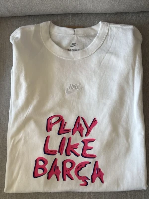 Vit Nike Barça t-shirt med tryck - Säljer en vit t-shirt från Nike med texten 'PLAY LIKE BARÇA' i rosa och svart framtill. Diskret Nike-logga på bröstet och Barça-logga samt grafiskt tryck på ryggen. Klassisk rund hals och korta ärmar. Helt ny med prislapp på då passformen inte passade. 