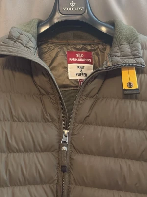 Olivgrön Cardigan Parajumpers - Säljer en olivgrön Cardigan från Parajumpers med stickade ärmar och quiltad front. Jackan har dragkedja, hög krage och den klassiska gula Parajumpers-detaljen vid halsen. Perfekt för kalla dagar och riktigt snygg till streetwear-stilen.