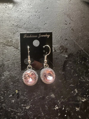 Silvriga örhängen med rosa sten - Snygga örhängen i silverfärg med rund, facetterad rosa sten i mitten. Stenen är omgiven av en dekorativ ram och örhängena har krokar för enkel användning. Perfekta för dig som vill ha något glittrigt och feminint.