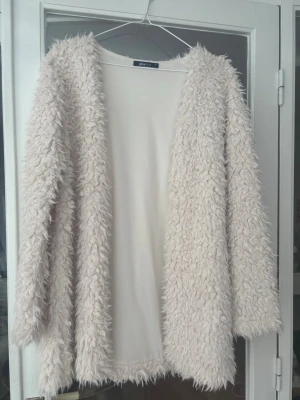 Beige fluffig cardiganjacka Gina Tricot - Fluffig teddyjacka i beige från Gina Tricot. Jackan har öppen front, lång ärm och är gjord i ett lurvigt, mjukt material som ger en cozy och trendig look. Perfekt för lager på lager under höst och vinter.