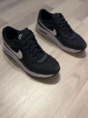  Nike Air Max  - Säljer ett par svarta Nike Air Max sneakers med vit sula och vit swoosh på sidan. Skorna har klassisk låg profil, snörning och synlig Air-enhet i hälen. Materialet är en mix av textil och syntet för en sportig look.