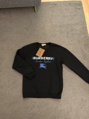 Svart Burberry sweatshirt med blått tryck - Svart sweatshirt från Burberry med blått och vitt tryck på bröstet där det står 'Burberry London, England' och en klassisk riddarlogga. Tröjan har rund halsringning och ribbade muddar. Perfekt för en stilren och trendig look.