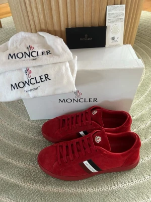 Röda Moncler sneakers i mocka - Snygga röda sneakers från Moncler i mjuk mocka med klassisk låg profil. Skorna har vita och svarta detaljer på sidan samt Moncler-logga på plösen. Perfekta för dig som vill sticka ut med stilrena och exklusiva sneakers.