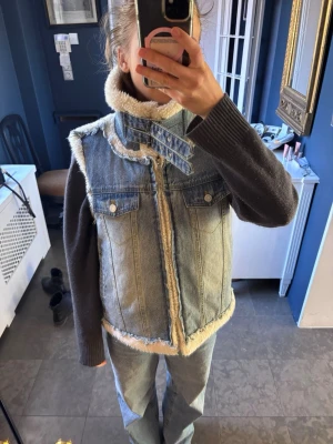 Blå jeansväst med fuskpälsdetaljer - Säljer en cool blå jeansväst med ljus fuskpäls längs krage, kanter och insida. Västen har två bröstfickor med knappar och dragkedja framtill. Den har en hög krage och råa kanter.