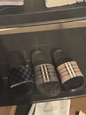 Rutiga slides i olika färger - Säljer trendiga slides med rutigt mönster i svart, grått och beige. Tofflorna har breda remmar och platt sula, perfekta för chill dagar. Materialet är syntet och designen är inspirerad av klassiska streetwear-vibbar.