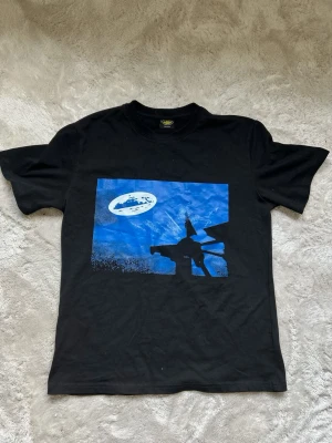 Svart Corteiz t-shirt med blått tryck - Cool svart t-shirt från Tank med ett stort blått grafiskt tryck på bröstet som föreställer Batman och ett rymdskepp. Klassisk passform och rund halsringning. Perfekt för dig som gillar unika motiv och streetwear.Använd en gång!