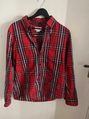 Röd rutig skjorta med knappar - Säljer en klassisk röd rutig skjorta med långa ärmar och knappar framtill. Skjortan har en avslappnad passform och är tillverkad i mjuk bomull, perfekt för en chill och stilren look. Passar lika bra till jeans som till kjol. Super fin och sitter skönt på, perfekt till alla väder. Super snygg röd skjorta. Bra passform och sitter snyggt på. Fin och skönt tyg. Perfekt på och snygg. Storlek 170 som är dam S. Typ aldrig använd. 