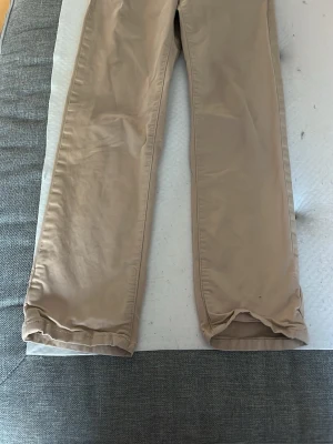Beige chinos från GANT - Snygga beige chinos från GANT med klassisk rak passform. Tillverkade i mjukt bomullstyg och har detaljer som blått innerfoder och randigt tyg på insidan av linningen. Perfekta för en clean och stilren look.