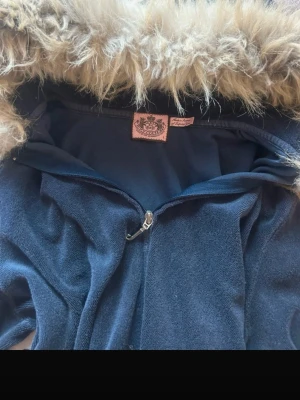 Juicy Couture jacka med fuskpäls - Mörkblå jacka från Juicy Couture med fluffig fuskpäls runt huvan och dragkedja framtill. Jackan har ett stort paljettmönster av en hund på ryggen och är tillverkad i mjukt material. Perfekt för dig som vill sticka ut med en unik och trendig look. Storlek L men passar S! 