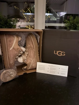 Ugg lowmel - Säljer ett par beige kängor från UGG med grova snören och fluffigt vitt ullfoder. Skorna har en robust sula och detaljer i mocka och textil. Perfekta för kalla dagar och riktigt snygga till jeans eller leggings.