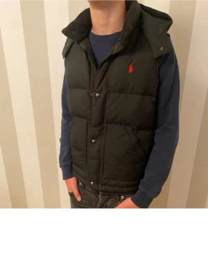 Svart dunväst från Ralph Lauren - Snygg svart dunväst från Ralph Lauren med röd logga på bröstet. Västen har avtagbar huva, tryckknappar och två stora fickor framtill. Perfekt för lager-på-lager och riktigt skön under kyliga dagar. Klassisk design som funkar till det mesta.