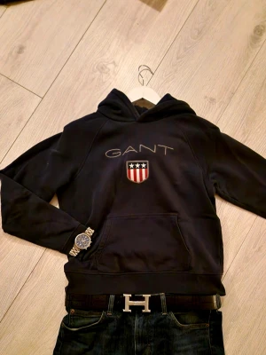 GANT Hoodie Mörkblå - Snygg mörkblå hoodie från GANT med broderad logga och ett sköldmärke med stjärnor och ränder på bröstet. Klassisk känguruficka framtill och huva. Perfekt för en avslappnad och stilren look. Fint skick, aldrig använd då den var för liten vid köp. Kolla gärna in mina andra annonser, ha det🤟