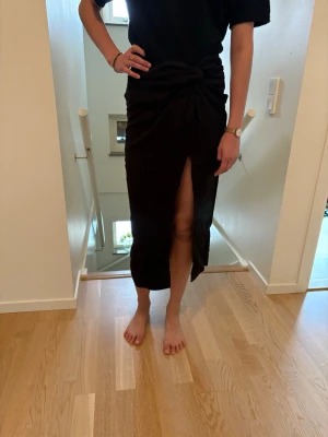 Svart långkjol med knyt och slits - Trendig svart långkjol med knyt i midjan och hög slits framtill. Kjolen har en elegant och enkel design som passar perfekt till många olika stilar. Materialet känns mjukt och följsamt.