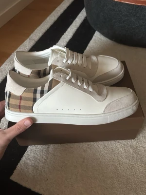 Burberry skor  - Snygga vita sneakers från Burberry med klassiskt beige, svart och rött rutmönster på hälen. Skorna har vita skosnören, detaljer i mocka och slät läderyta. Perfekta för dig som vill ha en clean look med lyxig touch.