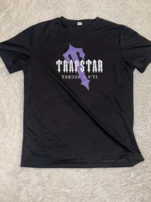 Trapstar svart t-shirt med tryck - Snygg svart t-shirt från Trapstar med stort lila och vitt tryck på bröstet där det står 'Trapstar It's a Secret'. T-shirten har rund hals och korta ärmar, perfekt för en streetwear-look. Materialet känns mjukt och bekvämt.