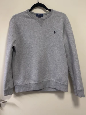 Grå sweatshirt från Polo Ralph Lauren - Säljer en stilren grå sweatshirt från Polo Ralph Lauren med rund halsringning och den klassiska mörkblå loggan broderad på bröstet. Tröjan har ribbade muddar vid ärmslut och nederkant, perfekt för en avslappnad och snygg look.