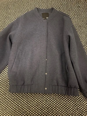 Mörkblå bomberjacka från 157 - Snygg mörkblå bomberjacka från 157 med minimalistisk design. Jackan har tryckknappar framtill, två snedställda fickor och ribbade muddar i nederkant. Materialet är mjukt och känns lite ulligt, med slätt foder på insidan. Perfekt för dig som gillar stilren och enkel look.