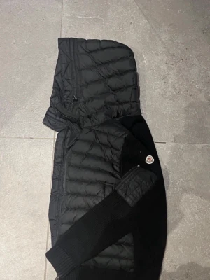 Svart pufferjacka från Moncler - Snygg svart pufferjacka från Moncler med stickade ärmar och quiltad kropp. Jackan har huva, dragkedja och Moncler-logga på ärmen. Perfekt för kalla dagar med sin kombination av dun och stickat material.