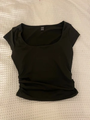Svart topp med rund hals från SHEIN - En svart, figurnära topp från SHEIN med korta ärmar och rundad, djup halsringning. Toppens material känns mjukt och stretchigt, perfekt för en stilren look. Passar dig som gillar enkla och snygga basplagg.
