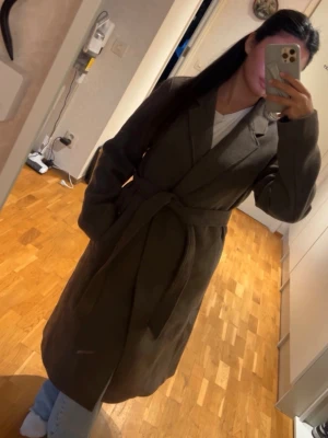 Mörkgrön lång kappa med knytskärp - Snygg mörkgrön kappa med klassisk krage och knytskärp i midjan. Kappan är lång och har en rak passform, perfekt för att ge en stilren look. Materialet ser ut att vara ullblandning och kappan har diskreta fickor på sidorna.