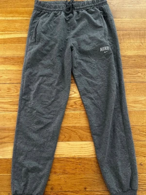 Grå Nike mjukisbyxor med logga - Säljer ett par grå mjukisbyxor från Nike med klassisk logga på vänster ben. Byxorna har elastisk midja med dragsko, sidfickor och ribbade benslut. Perfekt för chill dagar och sport. Materialet är mjukt och bekvämt.