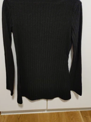 Svart ribbstickad tröja - En svart, långärmad ribbstickad tröja med smal passform och rund hals. Tröjan har diskreta vertikala ränder i stickningen och är tillverkad i ett mjukt, stretchigt material som sitter snyggt på kroppen. Perfekt basplagg för lager på lager. Storlek L 