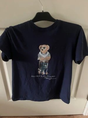Mörkblå Polo Bear t-shirt Ralph Lauren - Mörkblå t-shirt från Ralph Lauren med ikoniskt Polo Bear-tryck på bröstet. T-shirten har rund halsringning och korta ärmar. Materialet är mjukt bomullstyg och trycket visar en björn i randig tröja och byxor. Perfekt för en avslappnad och trendig look.