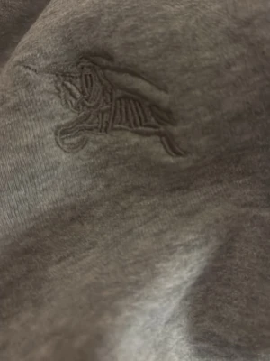 Grå sweatshirt från Burberry - Snygg grå sweatshirt från Burberry med det klassiska broderade ryttaremblemet på bröstet. Insidan är mjuk och fleecefodrad för extra komfort. Perfekt för dig som vill ha en stilren och exklusiv tröja med ikonisk Burberry-känsla.