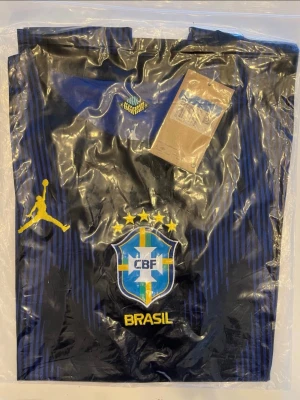 Helt ny! Brasilien 2026 tröja  - Helt ny💯Säljer en blå och svart träningströja från Jordan i samarbete med Brasilien fotbollslandslag. Tröjan har gula detaljer, CBF-logga och Jordan-logga på bröstet samt diskreta ränder. Perfekt för fotbollsträning eller att bära till vardags. 
