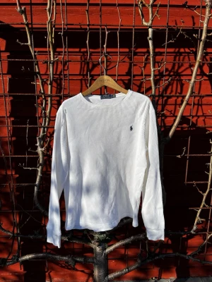 Vit långärmad tröja från Polo Ralph Lauren - Klassisk vit långärmad tröja från Polo Ralph Lauren med rund hals och diskret blå logga broderad på bröstet. Helt OANVÄND!!!!! Orginal storlek L men passar M. Tillverkad i ett mjukt, ribbat bomullstyg som ger en avslappnad och stilren look.Priset ej hugget i sten. Hör av er vid frågor eller funderingar! 