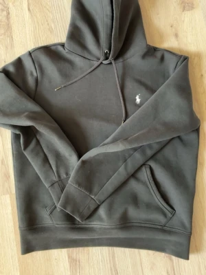 Svart hoodie från Polo Ralph Lauren - Snygg svart hoodie från Polo Ralph Lauren med klassisk huva och dragsko. Ikoniska loggan broderad på bröstet i vitt. Mjuk bomullsblandning och känguruficka framtill. Perfekt för en avslappnad och stilren look. Kom gärna med byten också!