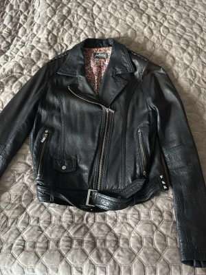 Svart skinnjacka från Jofama - Snygg svart skinnjacka från Jofama med klassisk bikerstil, sneddragna dragkedjor, bälte i midjan och nitar vid sidan. Jackan har blommigt innerfoder och flera fickor med dragkedja. Perfekt för en cool och tidlös look. säljer då den aldrig kommer till användning.