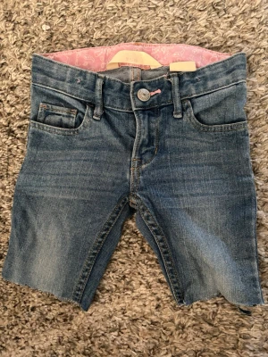 Blå jeansshorts med rå kant - Säljer ett par blå jeansshorts med rå, avklippt kant och klassisk femficksdesign. Shortsen har normal passform och midja, samt knapp och dragkedja framtill. Perfekta för sommaren och ger en avslappnad look.